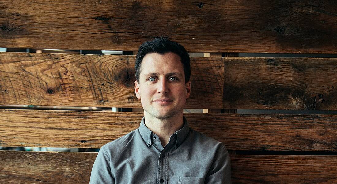 A young man in a casual grey shirt sits against a dark wooden wall.