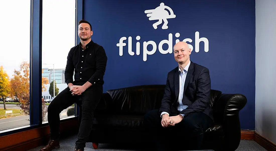 Two men in the Flipdish headquarters in Dublin. One man in a dark shirt and jeans is perched on the arm of a black leather sofa. The other, in a light shirt and dark suit, is seated on the other end.