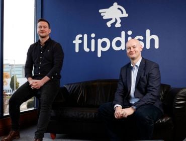 Two men in the Flipdish headquarters in Dublin. One man in a dark shirt and jeans is perched on the arm of a black leather sofa. The other, in a light shirt and dark suit, is seated on the other end.