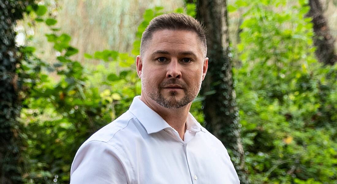 A man in a white shirt stands outdoors surrounded by trees and greenery. He is smiling at the camera.