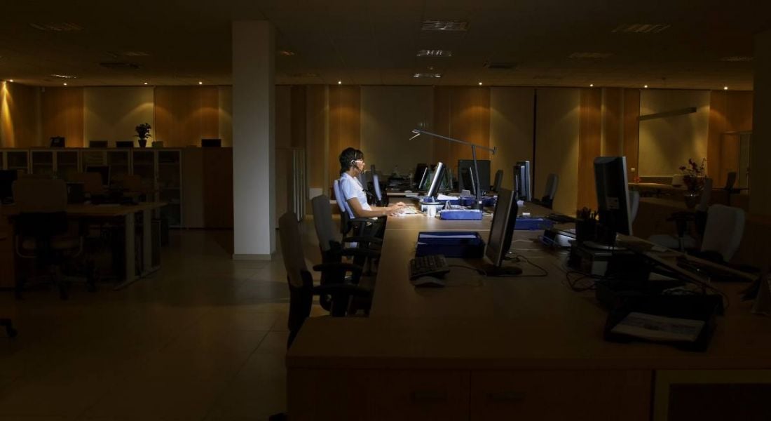 A young woman working at desk in a dark office alone. She is surrounded by empty desks, symbolising the great resignation.