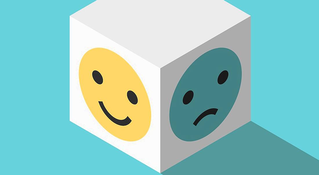 A 3D white cube on a blue background. One side shows a happy yellow emoticon, while the other side shows a sad blue emoticon to symbolise imposter syndrome.