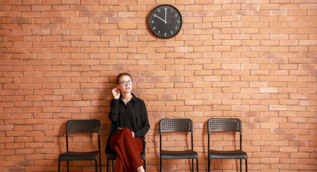 A young woman sits on one of four black chairs against a brick wall with a clock over her head. She is waiting for a job interview.