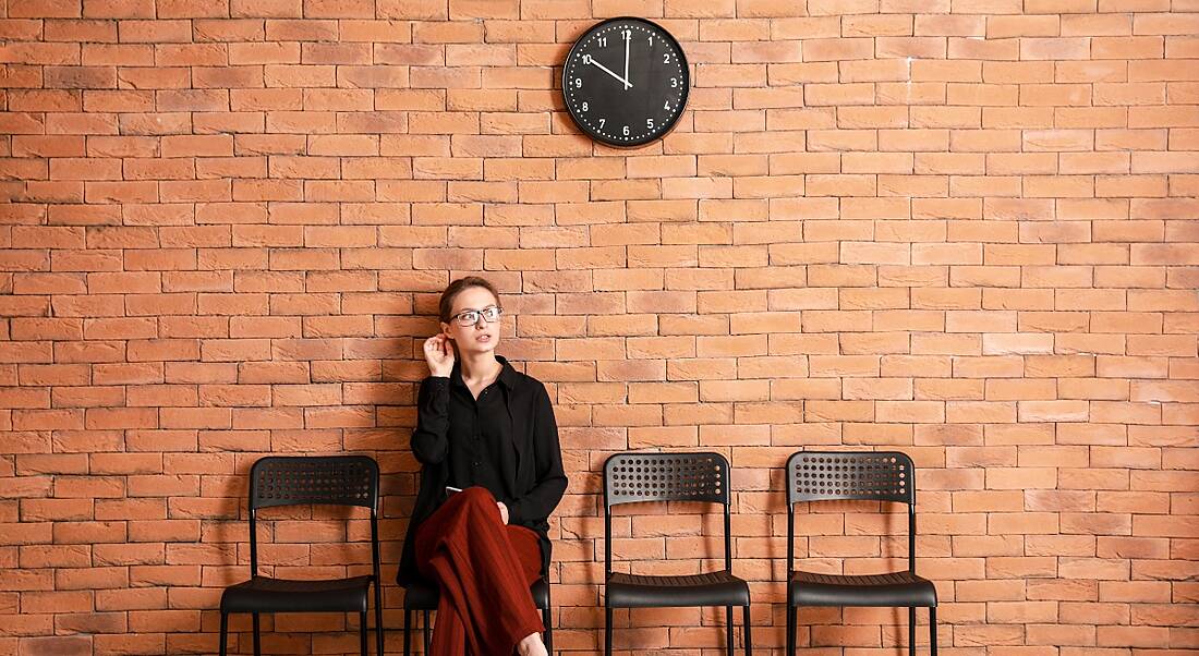 A young woman sits on one of four black chairs against a brick wall with a clock over her head. She is waiting for a job interview.