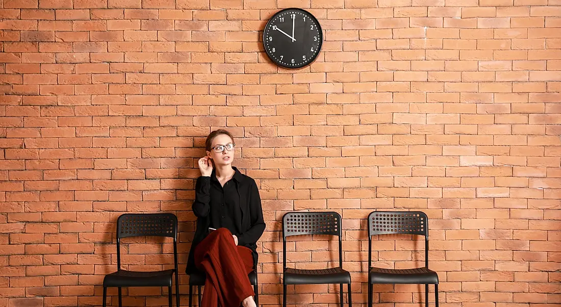 A young woman sits on one of four black chairs against a brick wall with a clock over her head. She is waiting for a job interview.