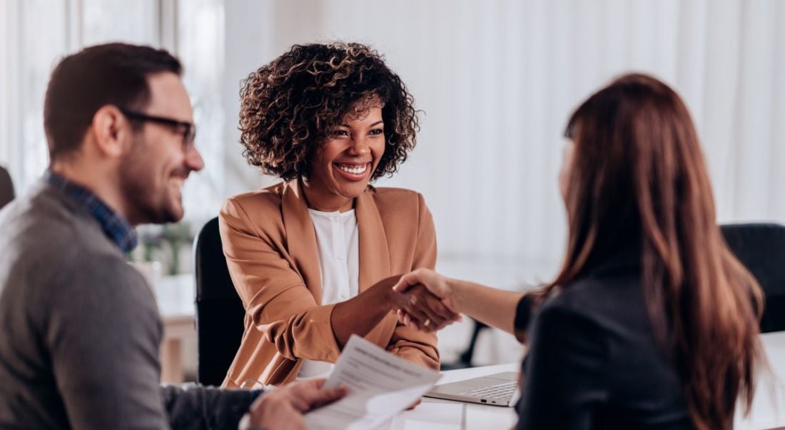 Two women shake hands at a job interview while a man sits beside them holding a CV.