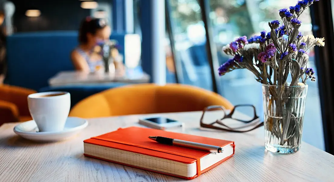 An orange notebook sits on a table with a pen resting on top. Beside it is a small vase of purple flowers, a cup of coffee, a pair of glasses and a phone.