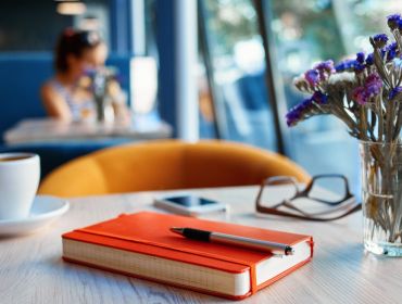 An orange notebook sits on a table with a pen resting on top. Beside it is a small vase of purple flowers, a cup of coffee, a pair of glasses and a phone.