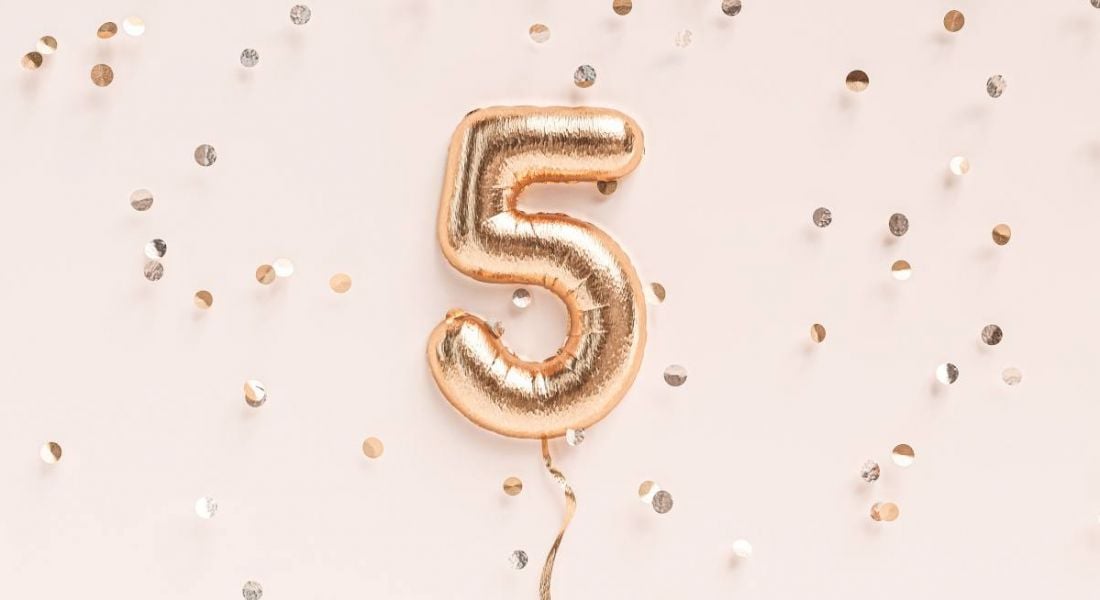 A rose gold foil balloon in the shape of a number five against a pale pink background surrounded by gold confetti.