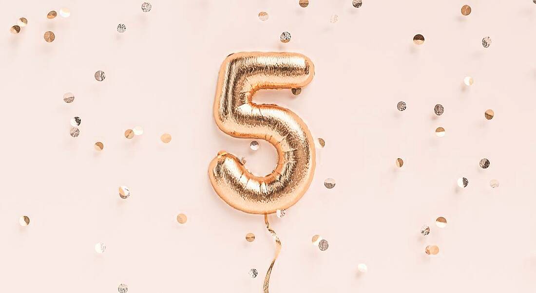 A rose gold foil balloon in the shape of a number five against a pale pink background surrounded by gold confetti.