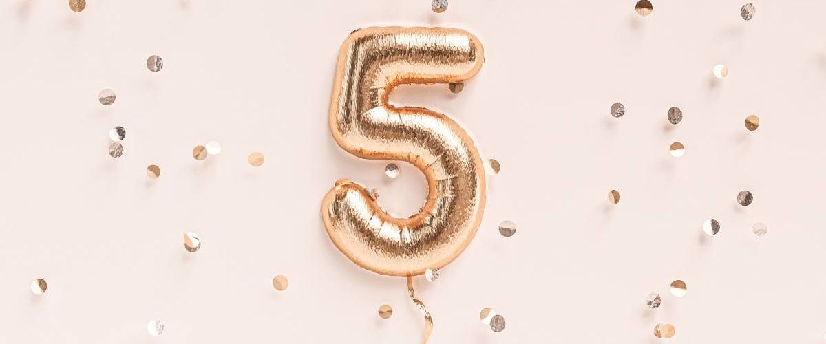 A rose gold foil balloon in the shape of a number five against a pale pink background surrounded by gold confetti.