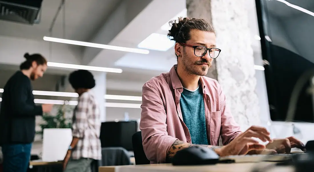 A man with glasses wearing a casual, open shirt over a t-shirt works at a computer in an office. Two co-workers are chatting in the background.