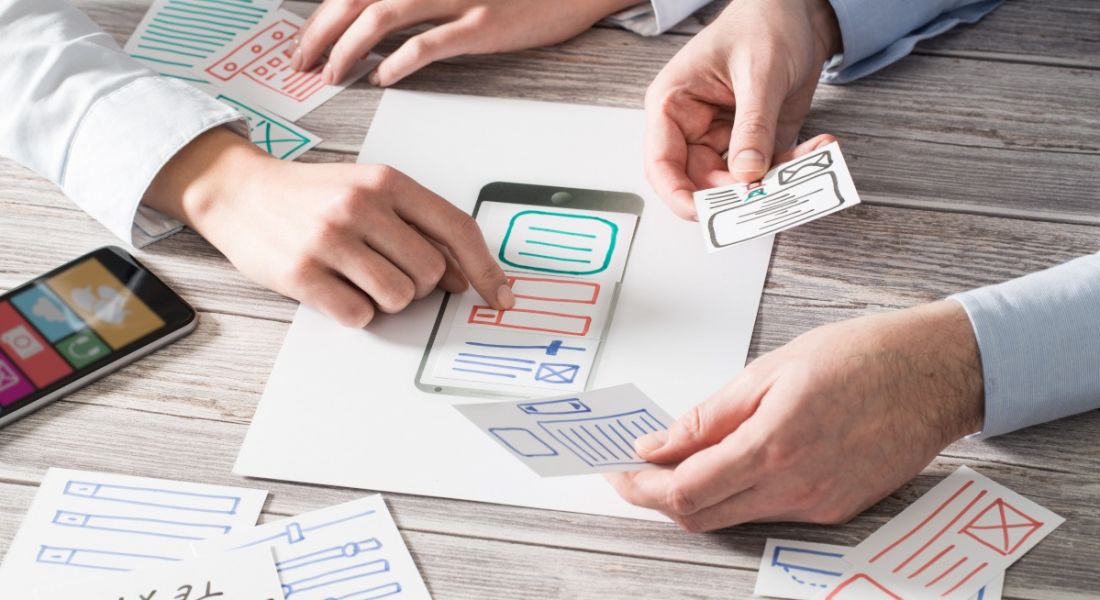 Two sets of hands look over pieces of paper with sketches of UX design ideas for a mobile app on them.