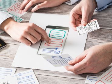 Two sets of hands look over pieces of paper with sketches of UX design ideas for a mobile app on them.