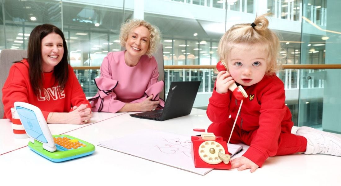 Two women sitting in an office smiling happily while a cute blonde haired toddler plays with a toy phone.