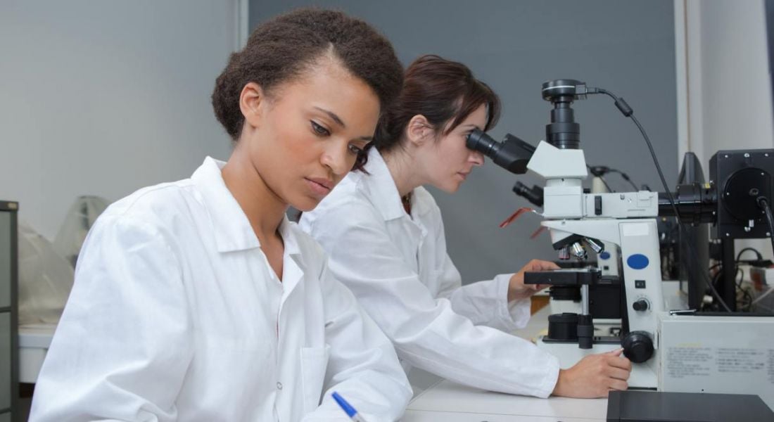 Two early-career women researchers dressed in lab coats working in a lab. One woman is taking notes while another looks at something through a microscope.