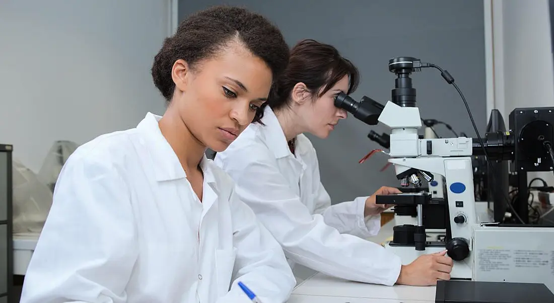 Two early-career women researchers dressed in lab coats working in a lab. One woman is taking notes while another looks at something through a microscope.