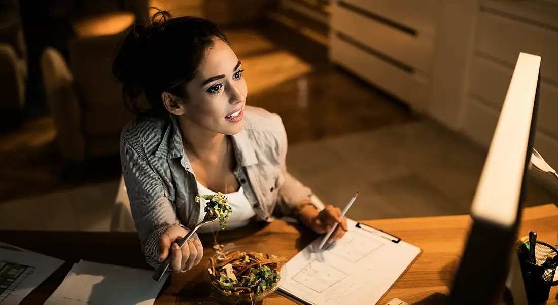 Woman working at a desk with a computer and a notebook in front of her. She is eating a salad and looking at the computer screen.