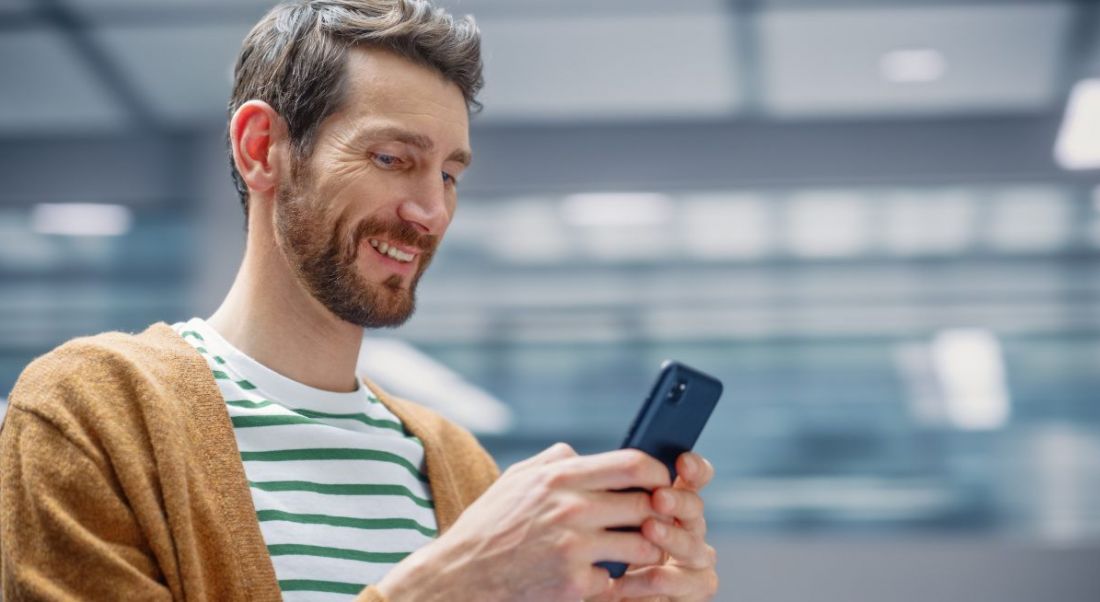 Man wearing striped green and white t-shirt and brown cardigan looking at his smartphone and smiling.