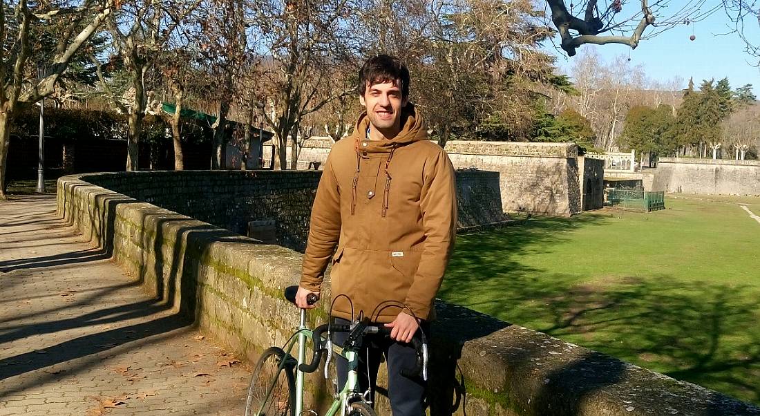 A young man in a tan jacket holds a bicycle outdoors while smiling at the camera.