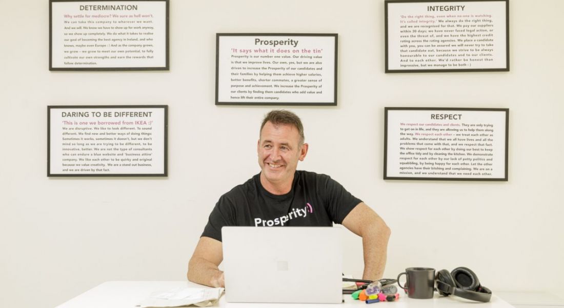 Gary Mullan of Prosperity Recruitment sitting in an office with cream coloured walls working at a laptop. Five framed pieces of text hang on the wall behind him.
