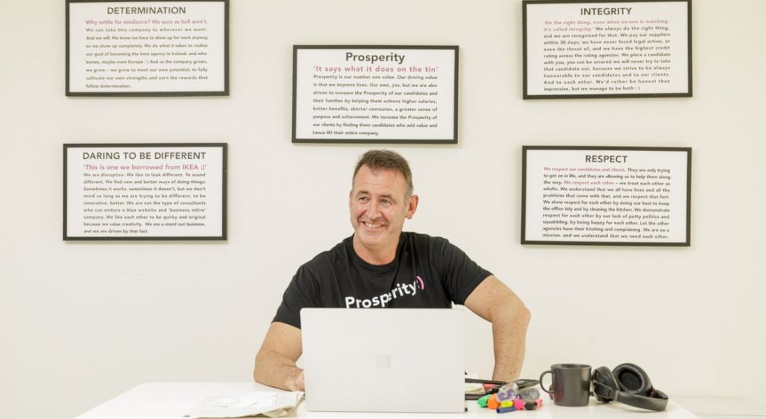 Gary Mullan of Prosperity Recruitment sitting in an office with cream coloured walls working at a laptop. Five framed pieces of text hang on the wall behind him.