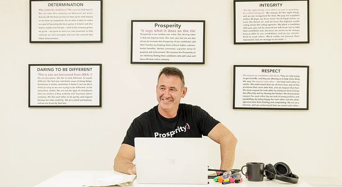 Gary Mullan of Prosperity Recruitment sitting in an office with cream coloured walls working at a laptop. Five framed pieces of text hang on the wall behind him.