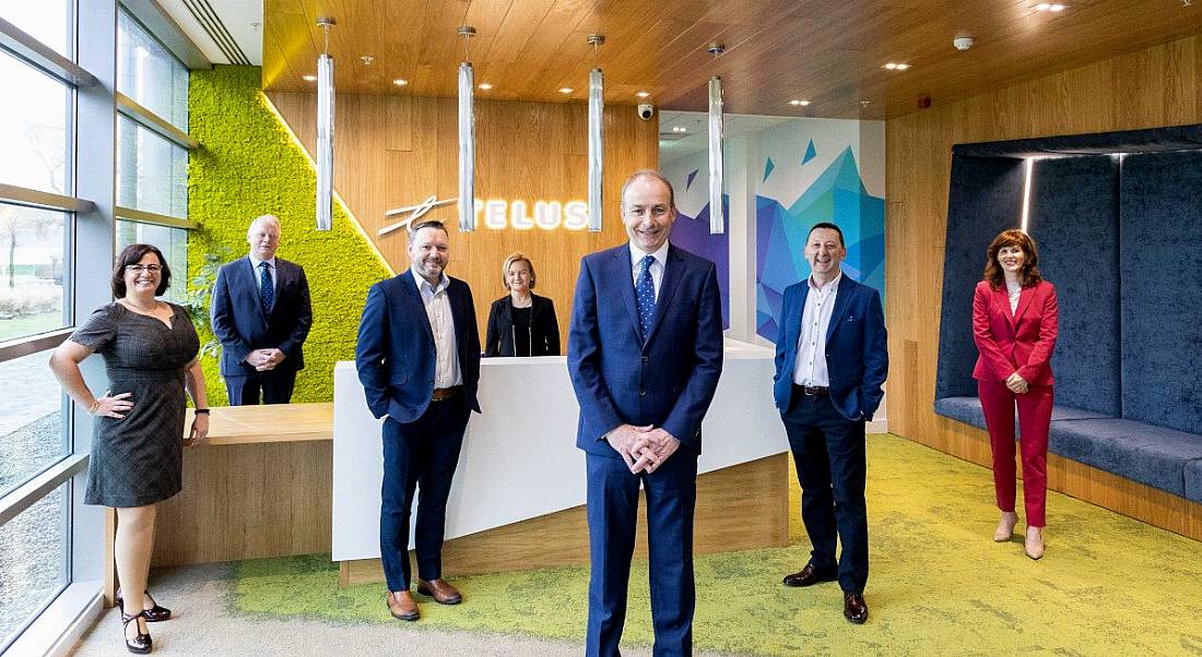 Four men and three women in smart business attire stand in a brightly lit, wood-panelled office reception. The word Telus is lit up behind the reception desk.