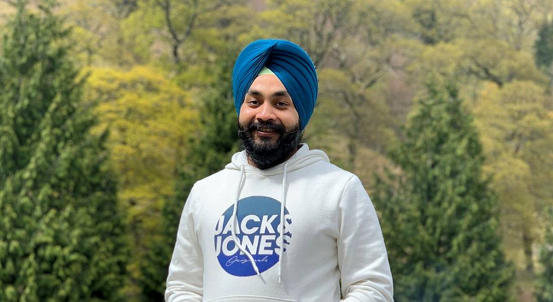 A man wearing a white hoodie smiles at the camera against the backdrop of a forest.