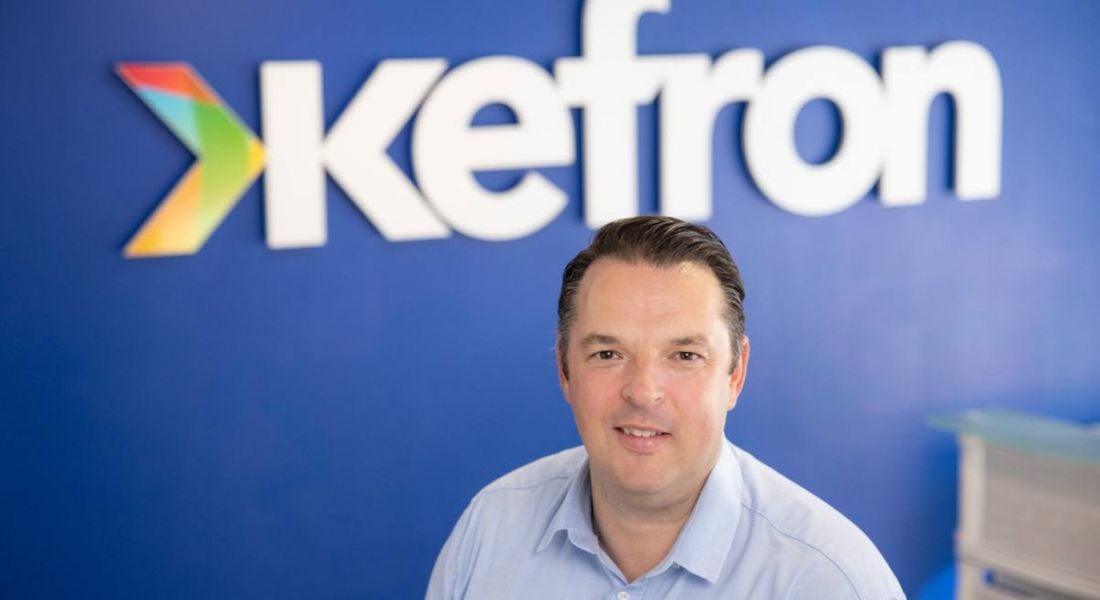 Paul Kearns, MD of Kefron sitting down in front of a blue wall with Kefron's logo.