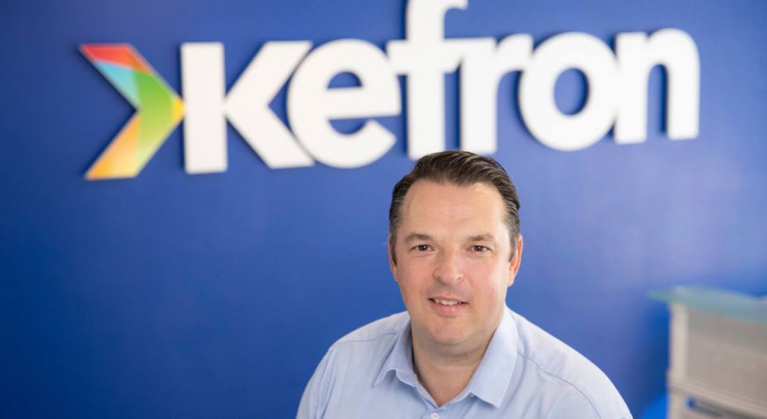 Paul Kearns, MD of Kefron sitting down in front of a blue wall with Kefron's logo.