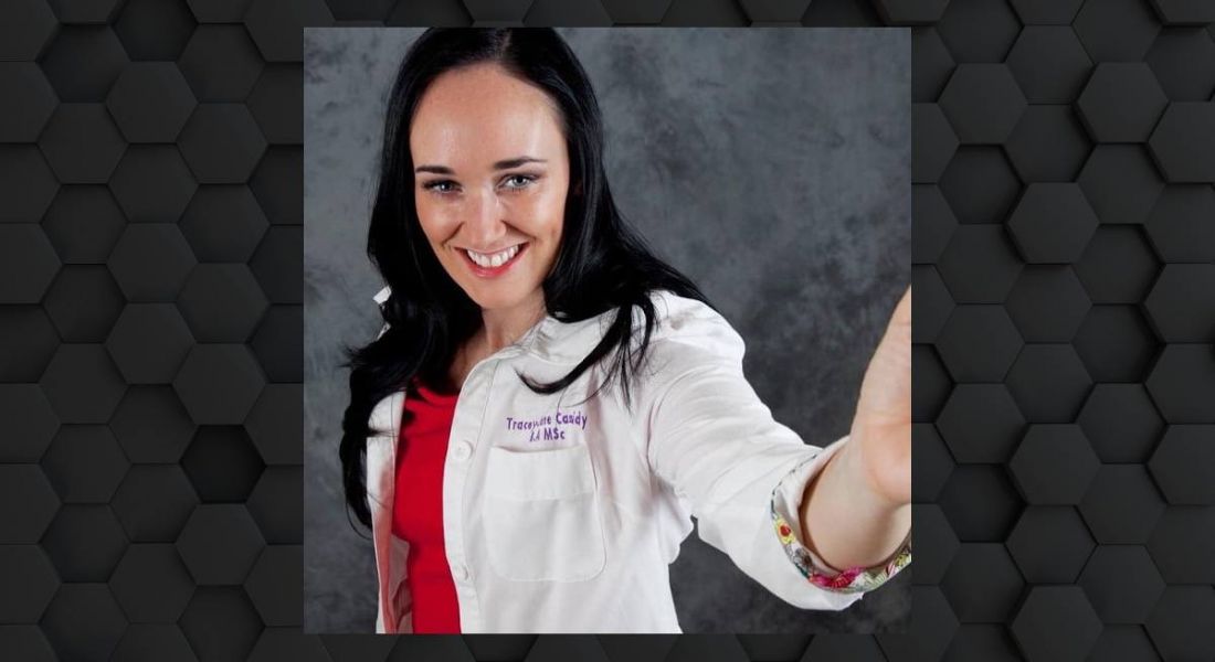 A woman with dark hair taking a selfie. She is wearing a white lab coat and smiling at the camera.