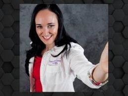 A woman with dark hair wearing a pink blazer and white blouse smiles at the camera against a dark wall. She is Susanne Jeffery from Accenture.