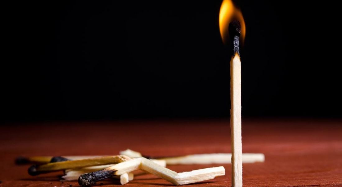 One burning match stands up on a wooden surface while several burnt broken matches lie beside it, symbolising tech worker burnout.