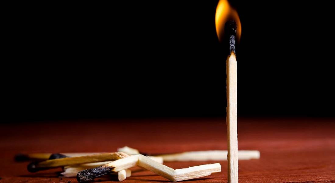 One burning match stands up on a wooden surface while several burnt broken matches lie beside it, symbolising tech worker burnout.