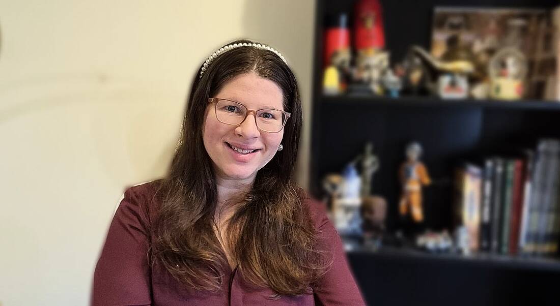 A dark-haired woman wearing a wine-coloured shirt smiles at the camera. Behind her is an out-of-focus black bookshelf with various items on it.