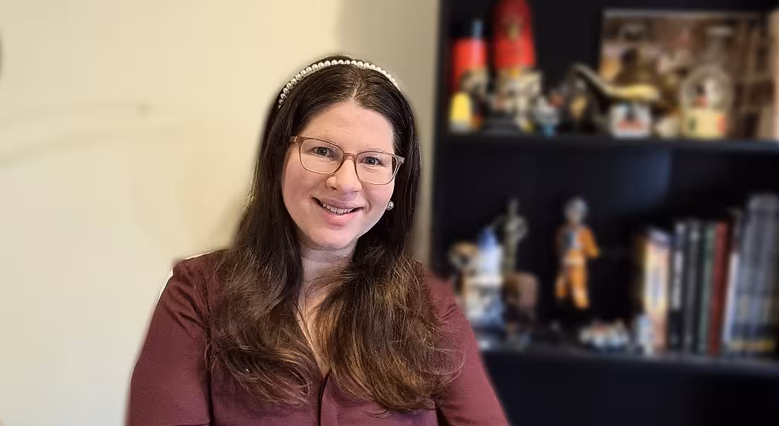 A dark-haired woman wearing a wine-coloured shirt smiles at the camera. Behind her is an out-of-focus black bookshelf with various items on it.