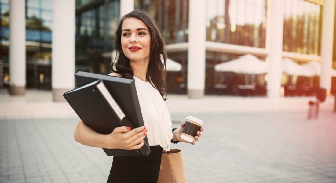 A young woman in business attire holding folders and a takeaway coffee cup on an urban area getting ready to return to the workplace.