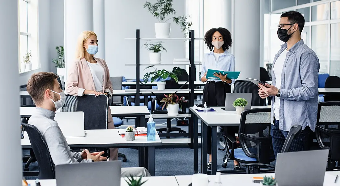 Two men and two women stand socially distanced in an office wearing masks, signifying a return to offices.