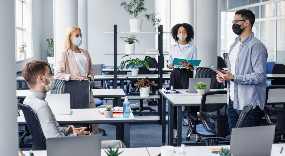 Two men and two women stand socially distanced in an office wearing masks, signifying a return to offices.