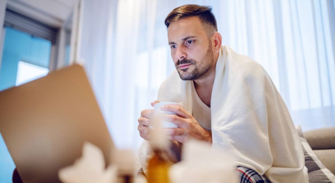 A man wrapped up in a blanket holding a cup of tea while staring a laptop, symbolising working on sick days.