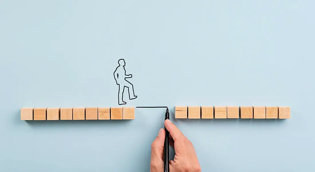 Wooden blocks are separated by a gap. An outline of a person is about to cross it while a large hand draws a line to bridge the gap. It symbolises the skills gap.