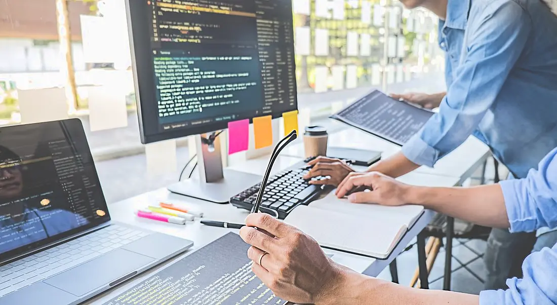 Two people at a desk with a laptop and a large screen, both of which have lines of code on them symbolising software development.