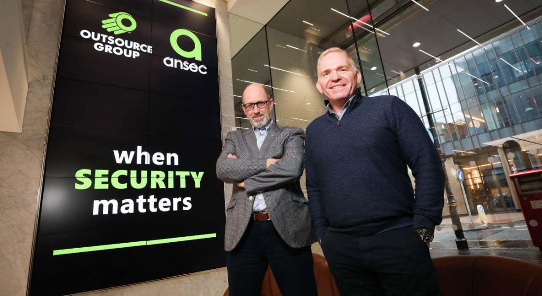 Peter Leitch and Terry Moore standing beside a large sign with ANSEC IA and Outsource Group logos on it in green writing following the merger of the Northern Ireland tech companies.