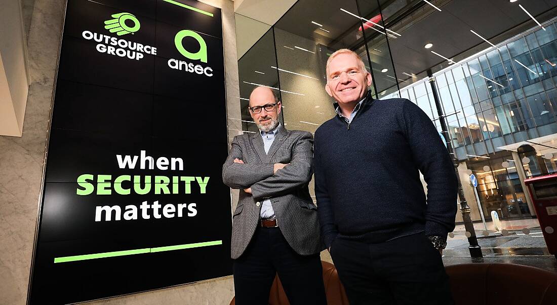 Peter Leitch and Terry Moore standing beside a large sign with ANSEC IA and Outsource Group logos on it in green writing following the merger of the Northern Ireland tech companies.