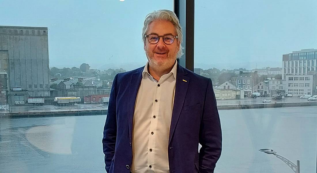 A man wearing a suit stands in front of a large open window overlooking a body of water. He is smiling at the camera.