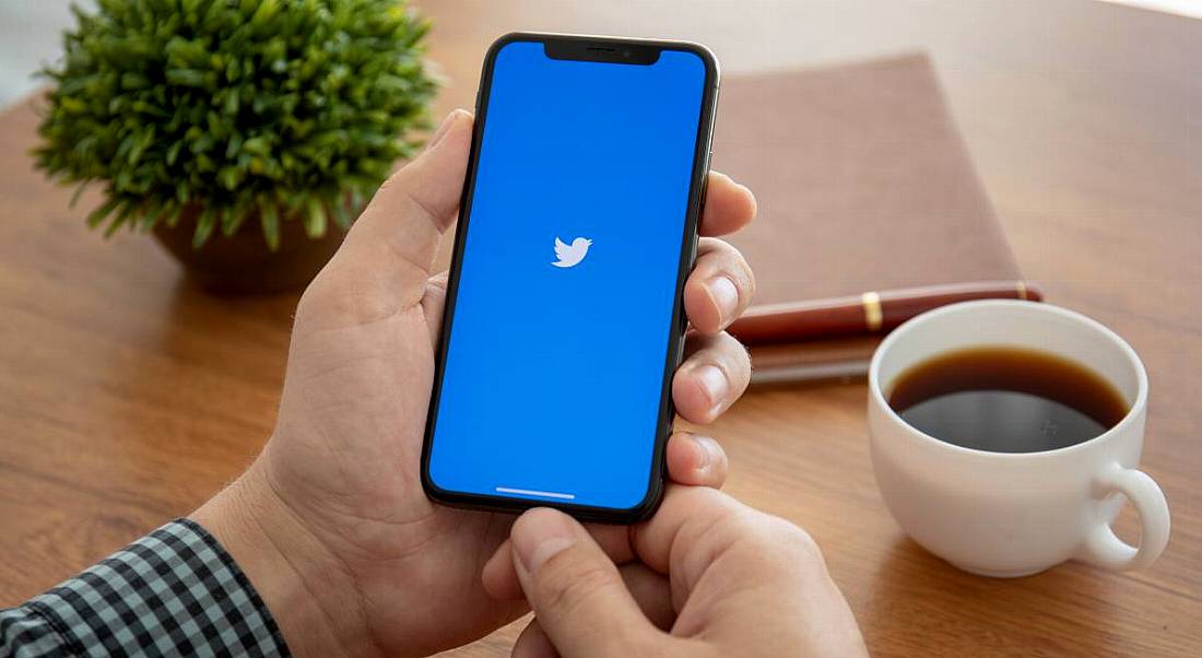 Man sitting at a wooden desk holding a smartphone with Twitter logo on it beside a cup of coffee, a notebook and a plant.