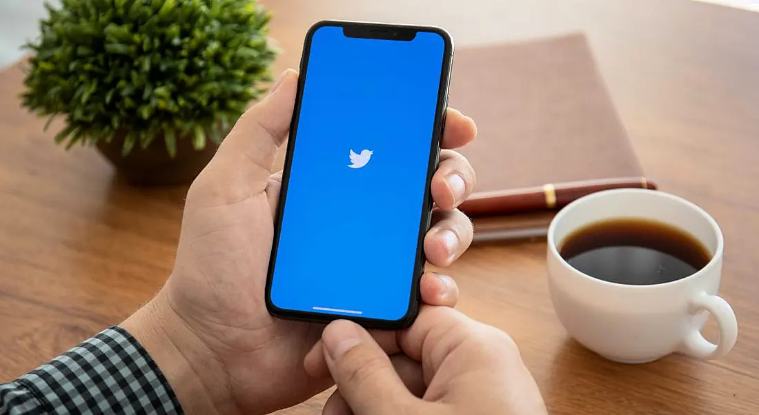 Man sitting at a wooden desk holding a smartphone with Twitter logo on it beside a cup of coffee, a notebook and a plant.