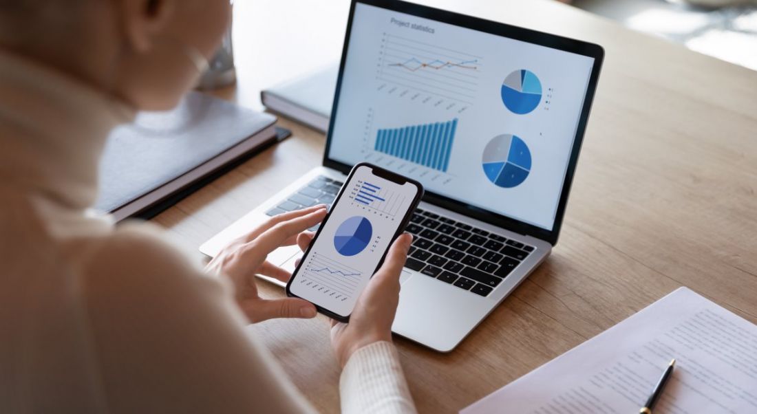 Woman sitting at a desk looking at statistics and pie charts on a laptop and on a smartphone.