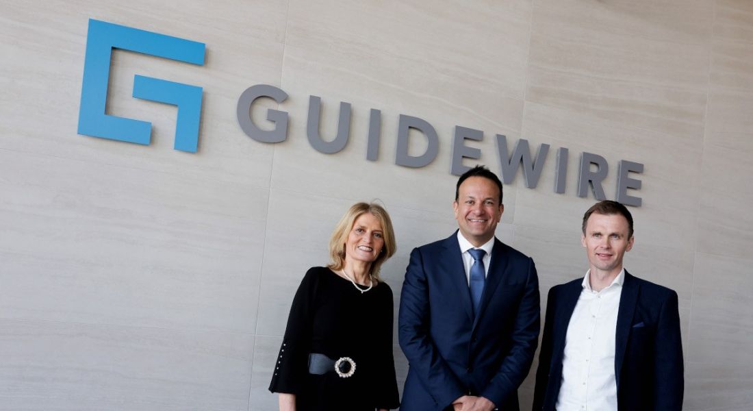 Three people stand in front of a wall that has the Guidewire logo on it.